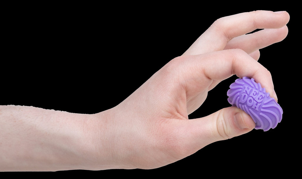 Teenie Singles M6 - A hand is squeezing a small, purple, textured ball between two fingers. The ball's surface features wavy ridges and is called a Teenie Ripples. The background is plain white.